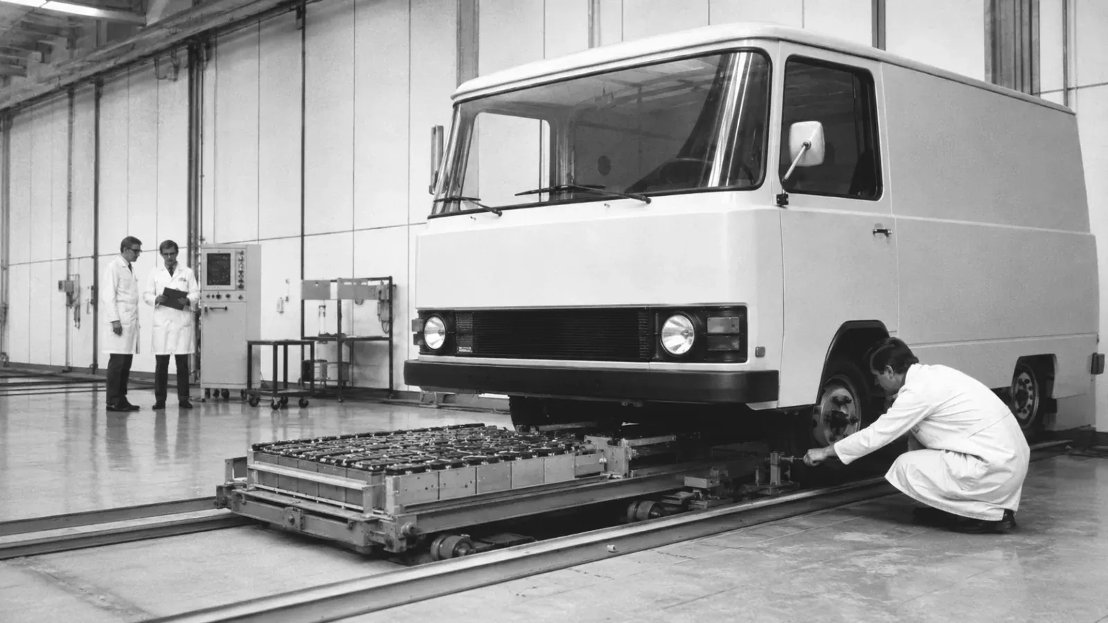Delivery van standing above a battery pallet on rails in a hall.